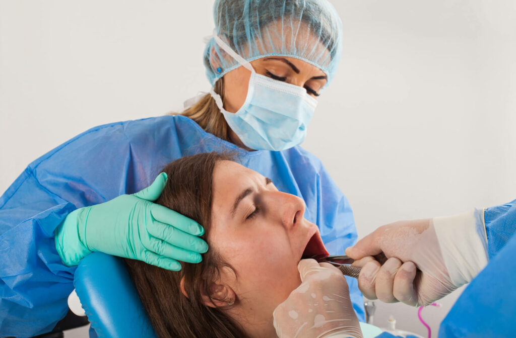 A dental surgeon and a dental assistant in a blue gown are removing the painful wisdom tooth of a female patient at the dental clinic.