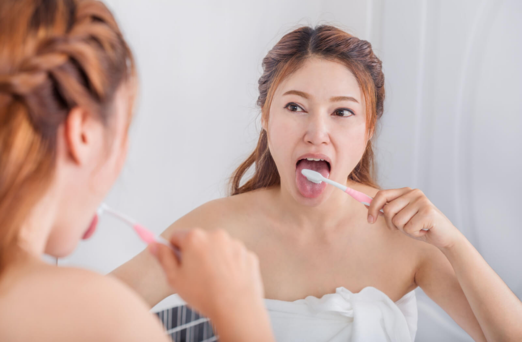 A young woman in the mirror is brushing her tongue to clean it.
