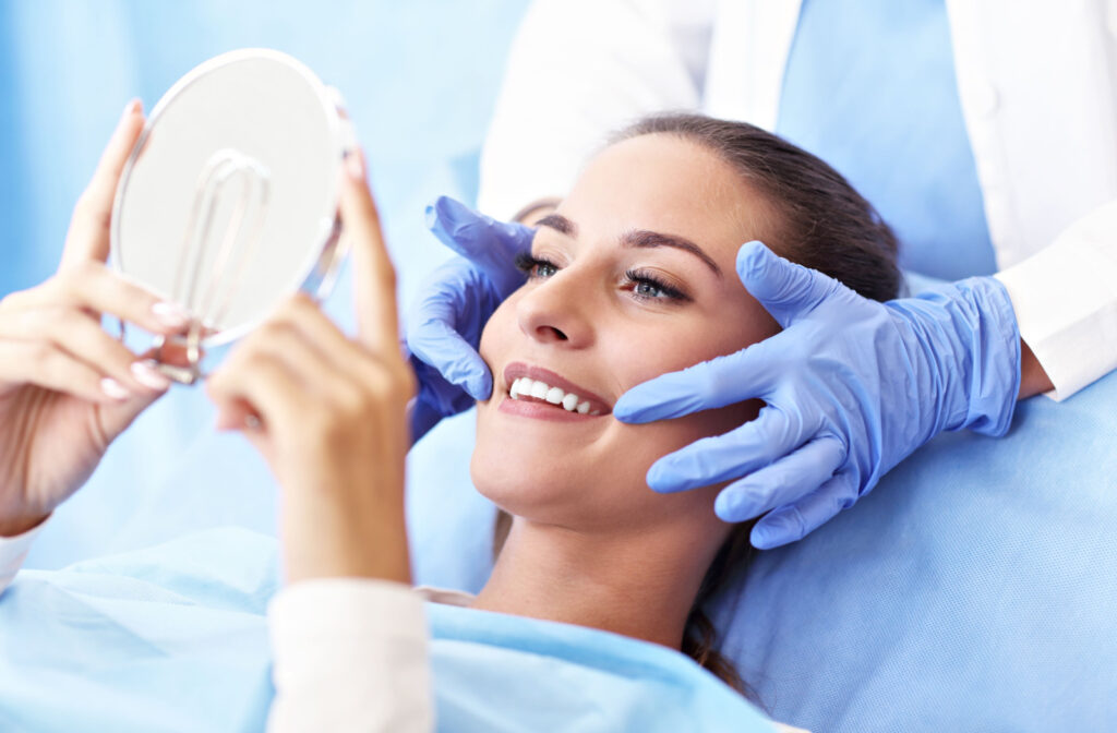 A woman lying in a dentist’s chair and smiling and holding a handheld mirror while her dentist gently holds the sides of her face.