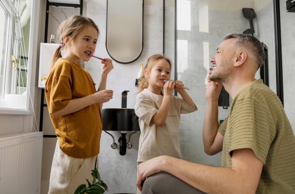 An adult and two children brush their teeth together in a modern bathroom, smiling and encouraging good oral hygiene habits.