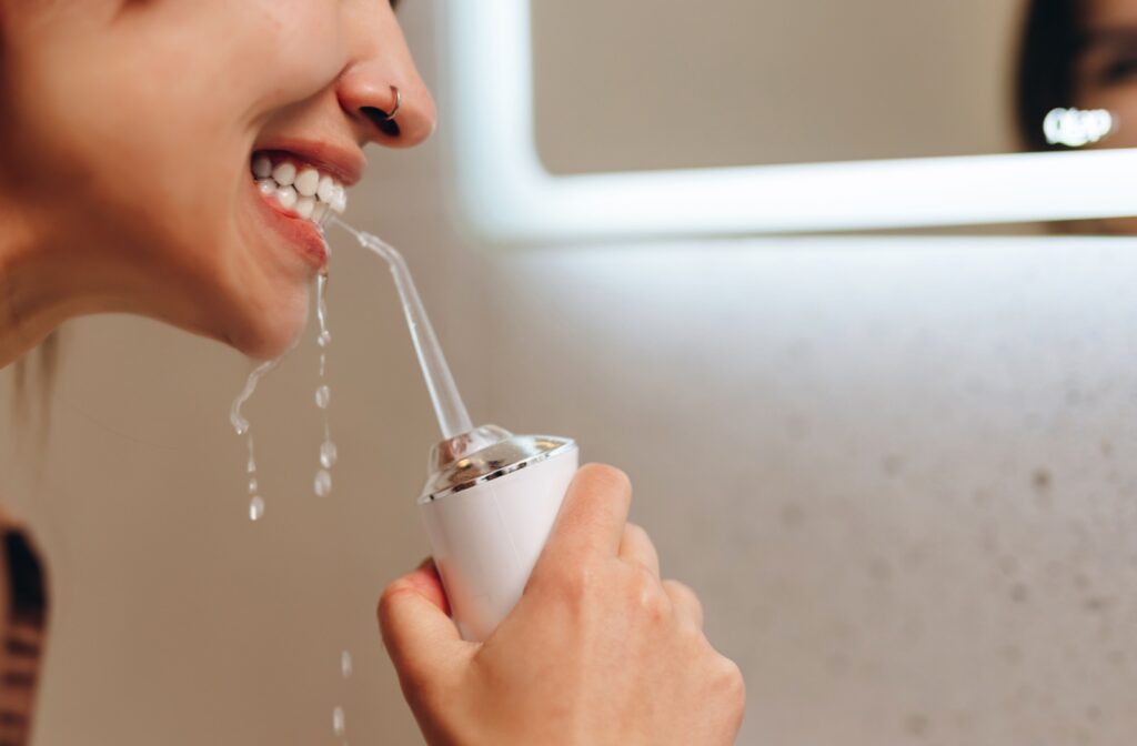 A woman using a water flosser in front of a bathroom mirror, with water stream aimed between teeth.