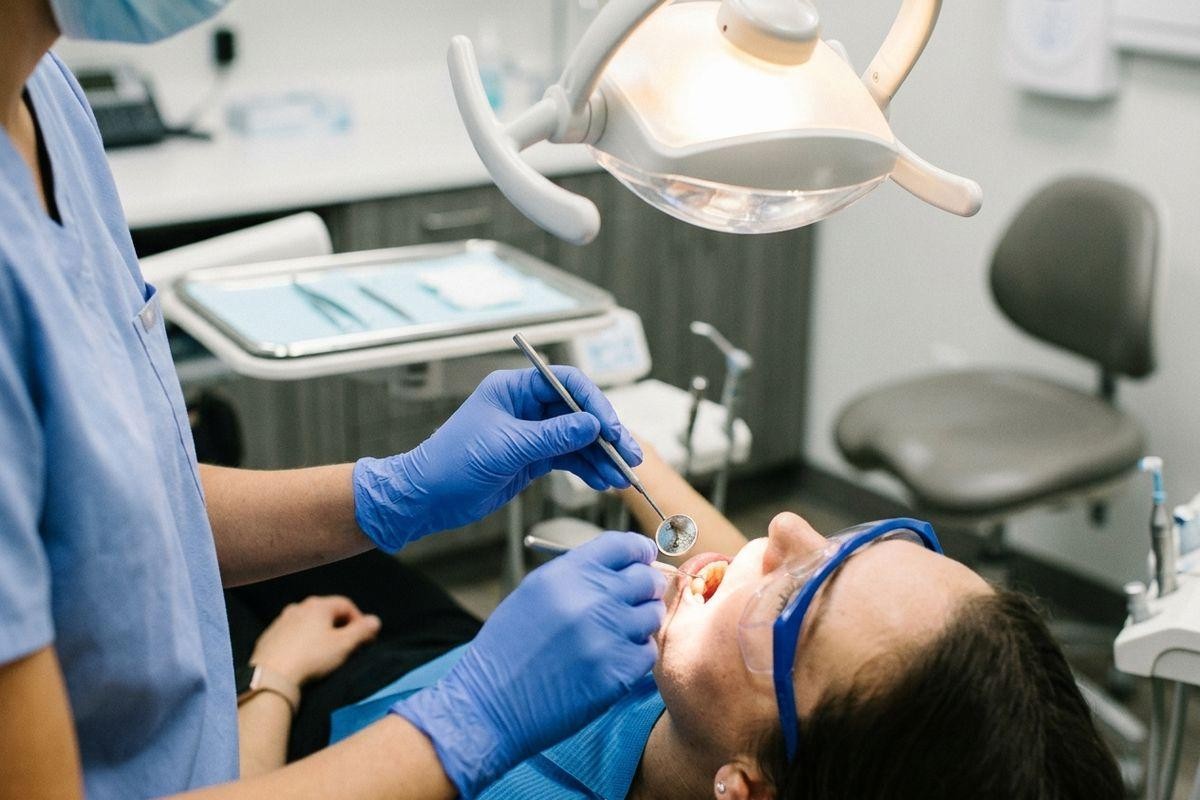 Dentist in blue scrubs and gloves examining a patient's open mouth with a dental mirror under a clinic lamp.